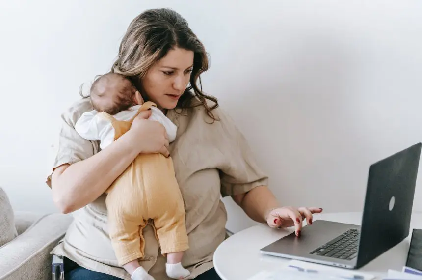 A woman holding a baby and looking at her laptop.