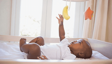 A baby laying in bed with a banana hanging from the ceiling.